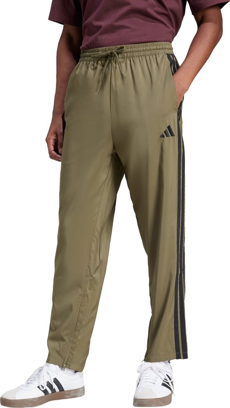 Man in een olijfgroene sportbroek met zwarte Adidas strepen, een koord in de taille en witte sneakers.