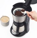 Hand voegt gemalen koffie toe aan filter van een Petra koffiezetapparaat, naast een mok koffie.