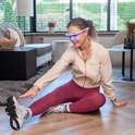 Vrouw met witte AYOlite lichttherapiebril in een woonkamer, zittend op de vloer tijdens het rekken.