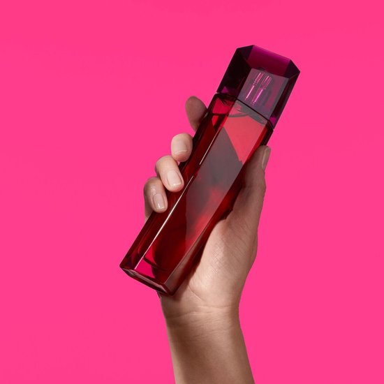 Hand houdt flesje Escada Magnetism Eau de Parfum in dieproze glas tegen roze achtergrond.
