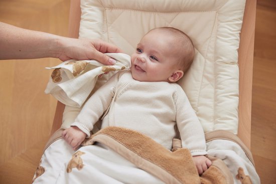 Bébé sourit pendant qu'une main tient la couverture blanche à imprimé ours en peluche. La couverture a un bord marron.