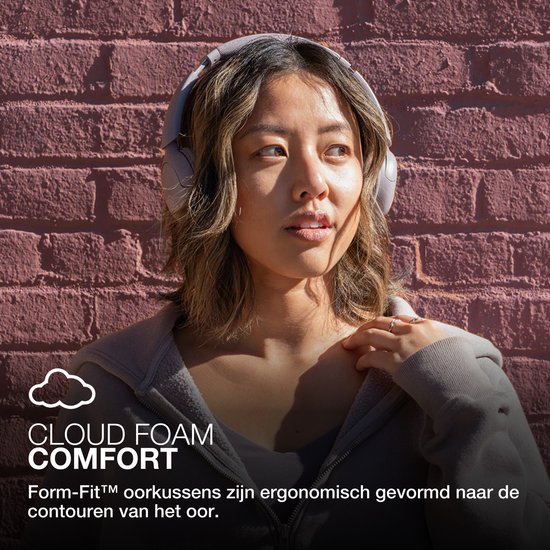 Vrouw in lichtgrijze koptelefoon bij bakstenen muur, met tekst 'CLOUD FOAM COMFORT'.