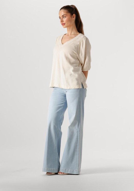 Model in een ecru blouse met V-hals, kapmouwtjes en geborduurde details op de mouwen. Draagt een lichte spijkerbroek.