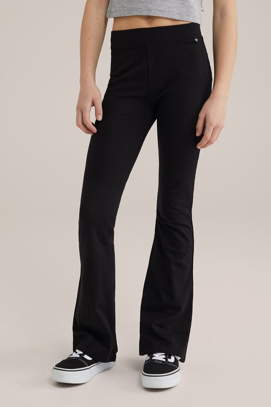 Model draagt een zwarte flared legging, met een klein wit sterretje op het tailleband-label. Zwarte Vans sneakers.