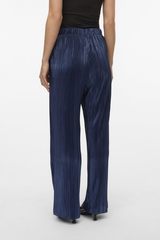 Model, achterkant, in een donkerblauwe, glanzende, wijde broek met plooien.