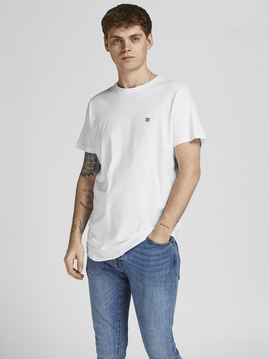 Man in wit t-shirt met kleine zwarte logoprint en jeans, hand in zak.