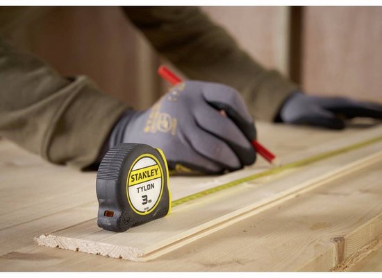 Man met grijze handschoenen markeert met potlood op houten planken, met Stanley rolbandmaat.