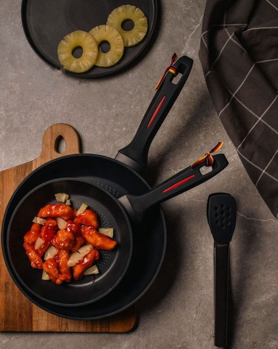 Poêle wok noire avec accents rouges et petits drapeaux allemands sur la poignée, remplie d'un plat de poulet et d'ananas. Des rondelles d'ananas sont à côté de la poêle.
