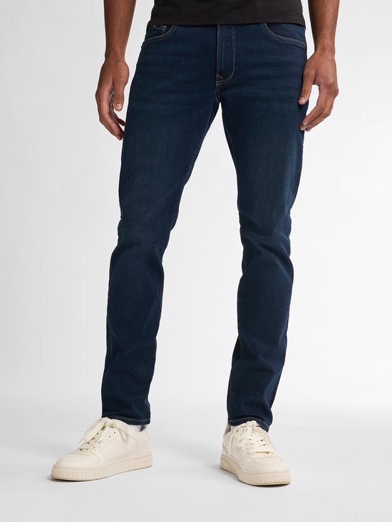 Voorkant van een man in een donkerblauwe slim fit spijkerbroek, staand op lichte sneakers.