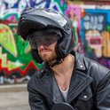 Man met baard en een matzwarte systeemhelm, met een leren jas, tegen een graffiti muur.