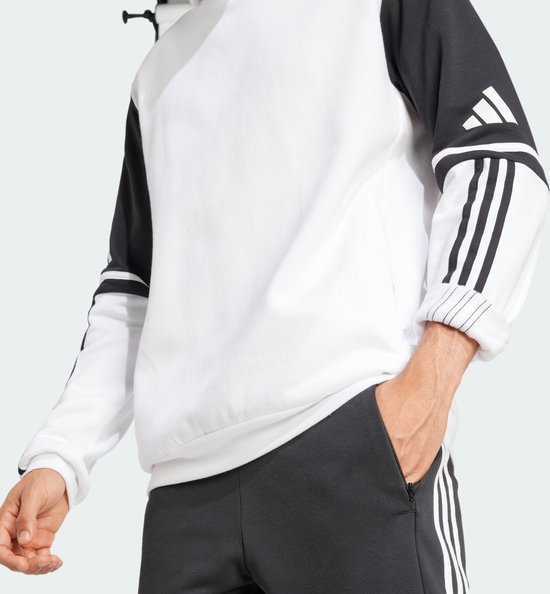 Man in een witte capuchontrui met zwarte mouwen met witte Adidas-strepen. Hij stopt zijn hand in zijn broekzak.