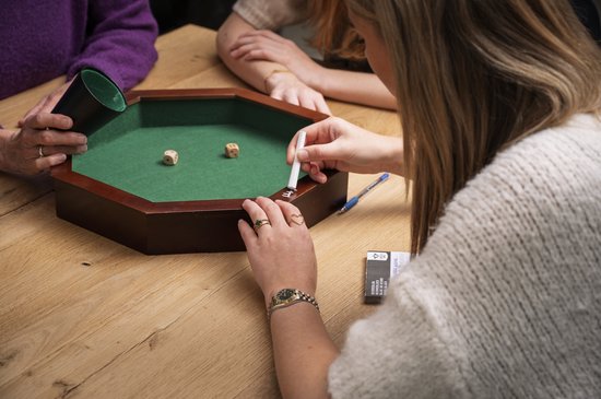 Drie mensen spelen een dobbelspel rond een achthoekige bak met groen vilt. Twee dobbelstenen liggen in de bak.