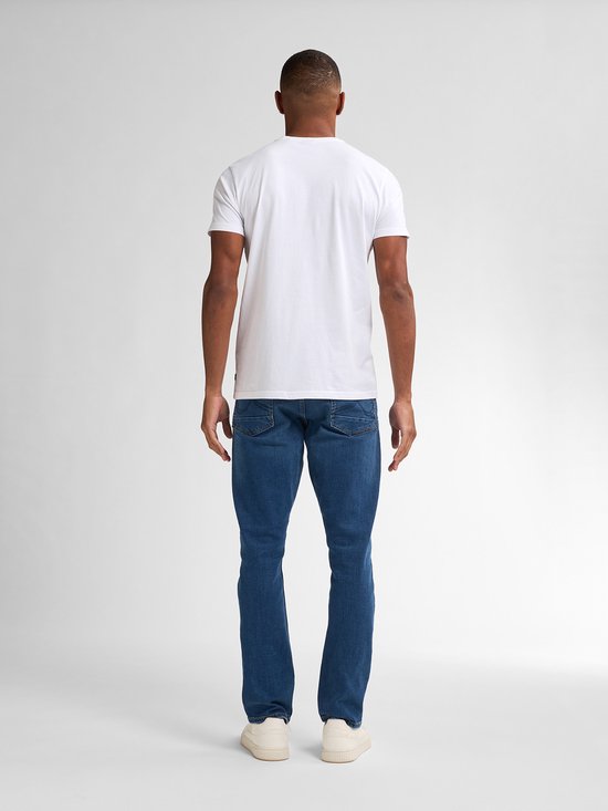 Man staat van achteren in een witte t-shirt en een blauwe spijkerbroek met rechte pasvorm, met witte sneakers.