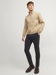 Man in een beige bomberjack en zwarte chino broek, staand.