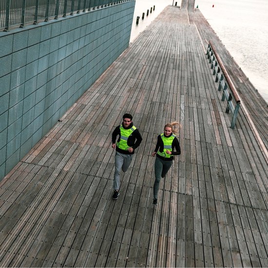 Man en vrouw joggend over een houten pier, beiden met een fluoriserend geel reflectiehesje.