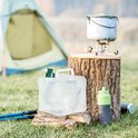 Kampeeruitrusting: opvouwbare jerrycan, wandelstokken, waterfles en kookpot op een houten stronk.