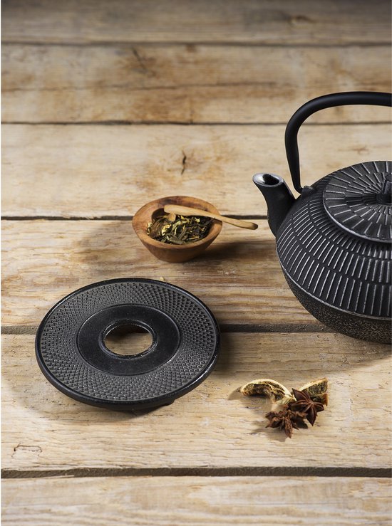 Zwarte gietijzeren onderzetter met puntig patroon naast een zwarte theepot op een houten tafel.
