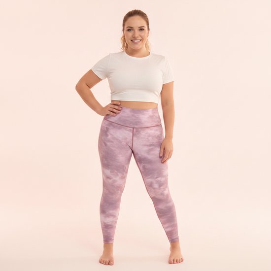 Vrouw in roze tie-dye legging en witte crop top, lachend naar de camera op een pastelroze achtergrond.