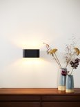 Zwarte ovale wandlamp op een dressoir met vazen, straalt warm licht uit.