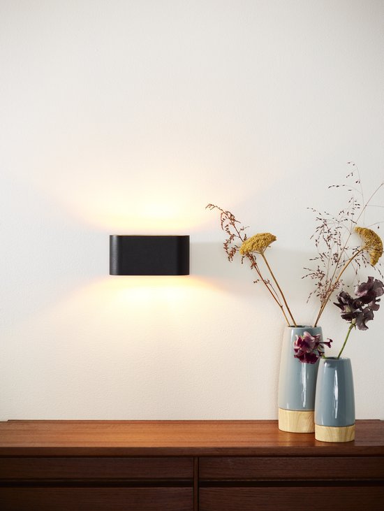 Zwarte ovale wandlamp op een dressoir met vazen, straalt warm licht uit.