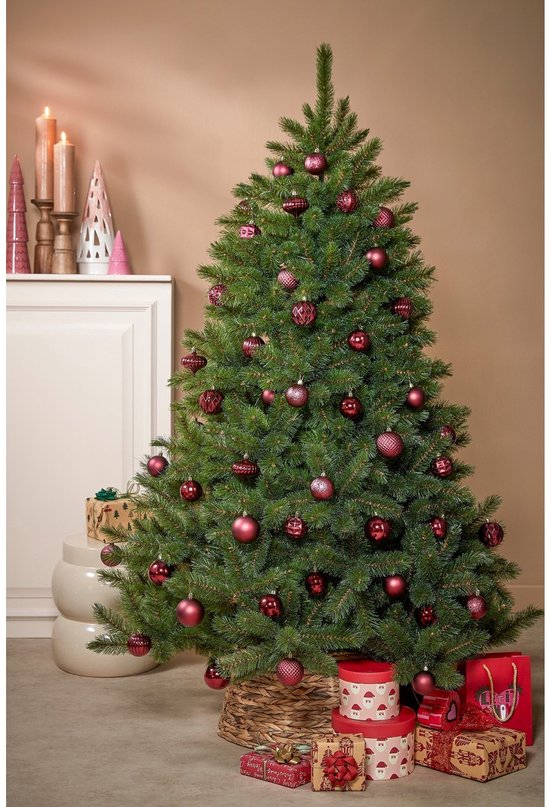 Een volle, groene kerstboom versierd met donkerrode kerstballen en staand in een rieten mand, met cadeaus eromheen.