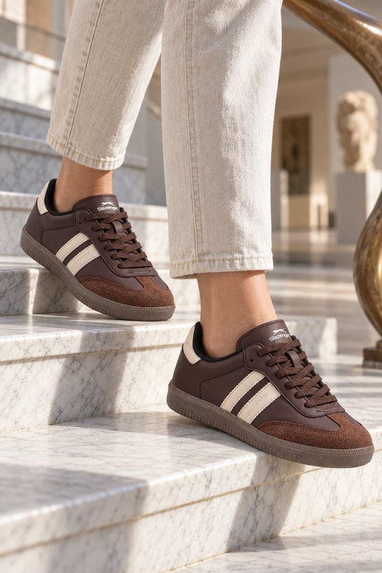 Bruine Slazenger sneakers met beige details, gedragen onder een lichte broek op een marmeren trap.