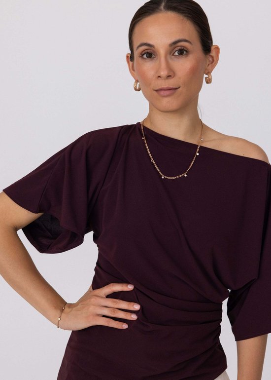 Model met gouden Purelei ketting met kleine steentjes en creolen. Draagt een paarse top met één mouw.