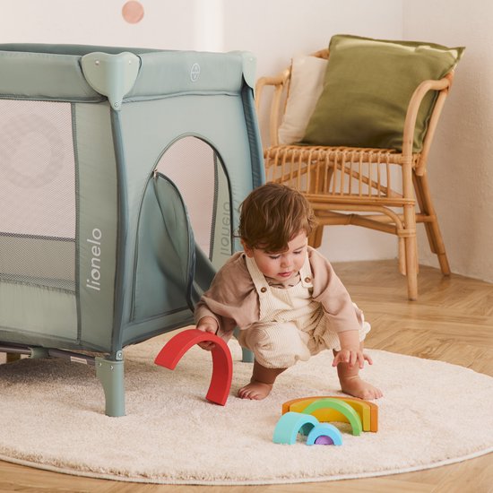 Peuter in beige kleding speelt met een regenboogvormige houten stapelaar naast een groen Lionelo reisbedje.