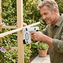 Man in een groen hemd gebruikt een wit-zwart nietpistool voor een houten lat in de tuin.