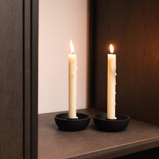 Twee brandende crèmekleurige dinerkaarsen in ronde, zwarte metalen kaarshouders op een donkere plank.
