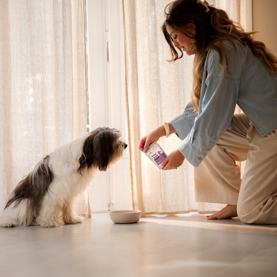 Vrouw knielt voor een hond en houdt een pakje 'bone broth eend' vast, bij een raam met gordijnen.