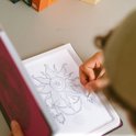 Hand die met een potlood een zonnemotief tekent op een wit tekenbord naast een roze Zen-notitieboek.