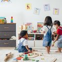 Grijze ladekast met 3 vakken, houten blad, houten handgrepen, in een kinderspeelkamer.