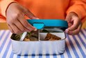 Kind sluit een zilverkleurige broodtrommel met een blauw deksel, gevuld met eten.