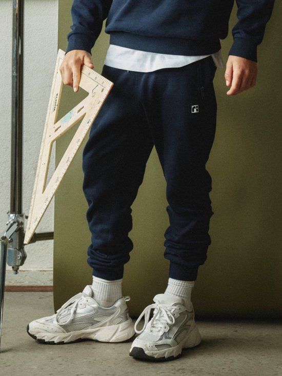 Man met donkerblauwe joggingbroek, witte sokken en sneakers, houdt een duimstok vast.