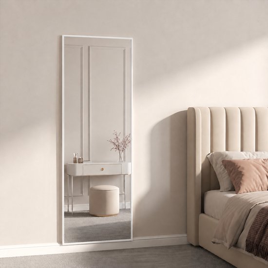 Een staande spiegel met witte lijst in een slaapkamer, naast een bed met beige hoofdbord.