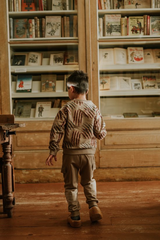 Een jongen kijkt naar een boekenkast, hij draagt een beige joggingbroek met cargo zakken en een trui met tijgerprint.