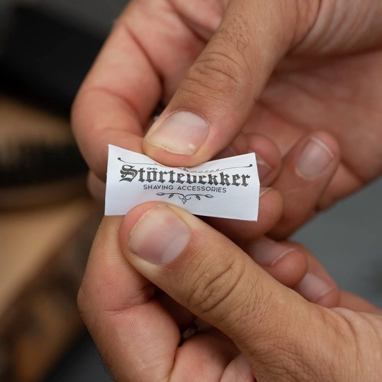 Handen houden een klein wit etiket met "Störtebekker SHAVING ACCESSORIES" in zwart schrift.