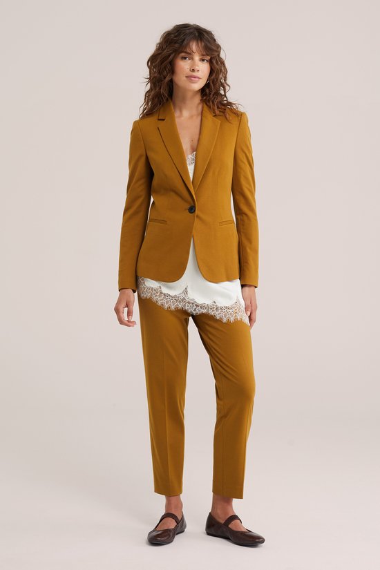 Model draagt een mosterdgele blazer met bijpassende broek, witte kanten top en bruine schoenen.