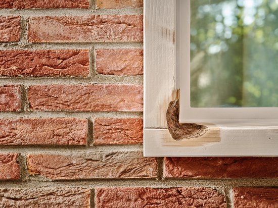 Een wit geschilderd houten kozijn met duidelijk houtrot op de hoek, naast een bakstenen muur.