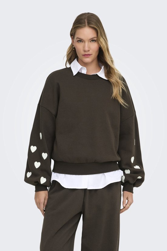 Donkerbruine sweater met witte hartjes op de mouwen, gedragen door een model met een witte blouse eronder.