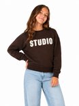 Meisje met bruine trui met 'STUDIO' in witte letters op de voorkant.