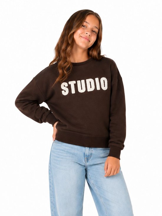 Meisje met bruine trui met 'STUDIO' in witte letters op de voorkant.