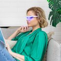 Vrouw met roze lichttherapiebril, leest een boek op een bank.