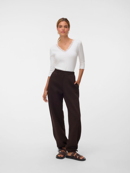 Model draagt een bruine joggingbroek met wijde pijpen en elastische tailleband, gecombineerd met een witte top.