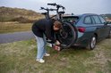 Vrouw in zwarte jas bevestigt een e-bike op een Cordo trekhaakfietsendrager achter een auto, in een duingebied.