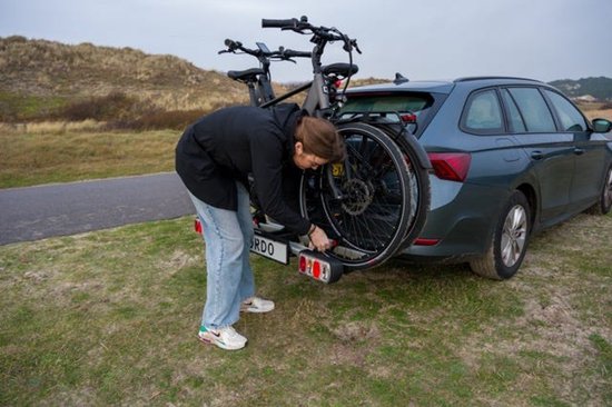Vrouw in zwarte jas bevestigt een e-bike op een Cordo trekhaakfietsendrager achter een auto, in een duingebied.