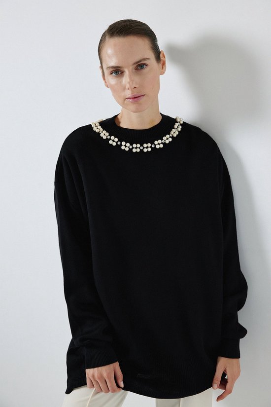 Model in zwarte trui met parelversiering aan de hals, gedragen met witte broek.