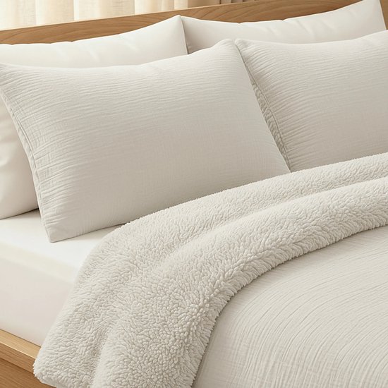 Twee beige kussenslopen van mousseline stof, met een subtiel geribbelde textuur. Een crème kleurige teddypluche deken ligt eroverheen gedrapeerd.