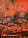 Vele papieren lampionnen en parasols, in wit en groen, hangen aan het plafond. Sfeervol oranje licht.